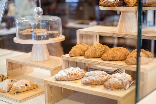 The pastry case, flaky croissants and other items can be seen inside a glass case.