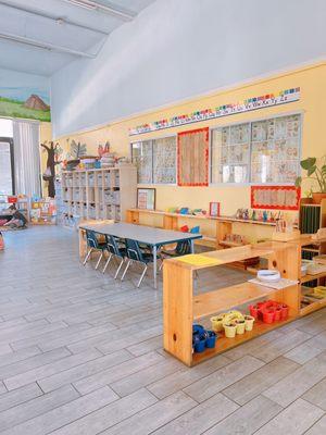 A spacious Montessori classroom. The room is filled with natural light, featuring large windows. There are various learning statio