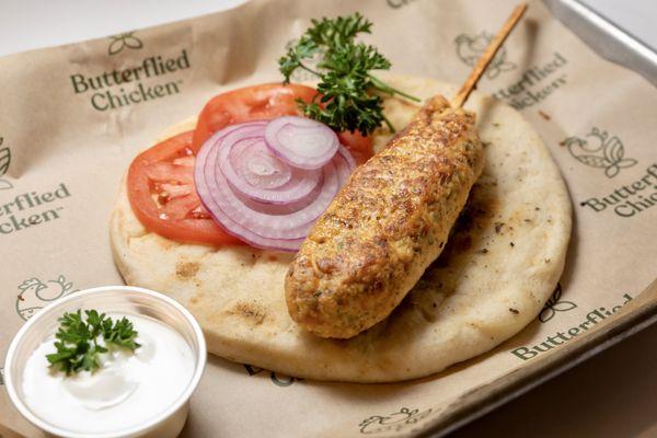 Chicken kebab on a stick, over a pita bread and garnished with onions and tomatoes with a side of mustard aioli sauce.