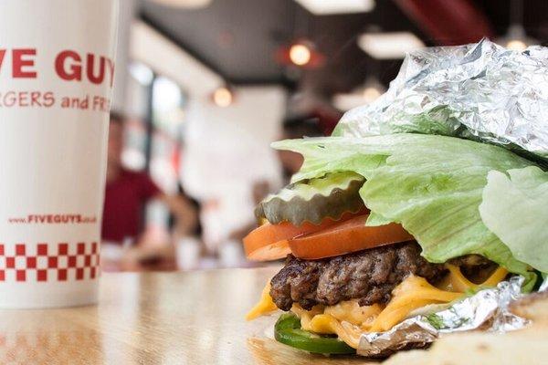 A close-up photograph of a Five Guys cheeseburger with pickles, tomatoes and jalapenos in a lettuce wrap.