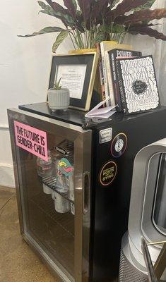 A mini-fridge housing refreshments. Queer and comedic stickers are pasted on the fridge, and there are plants and books on top.