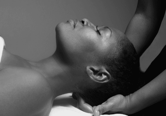 A Black woman is relaxing on a massage table. Her eyes are closed and her head is gently held by the hands of a Black massage therapist.