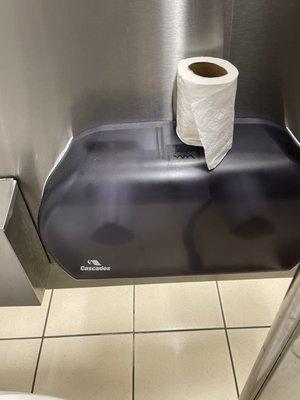 Toilet paper dispenser empty, toilet paper on top of dispenser, next stall.