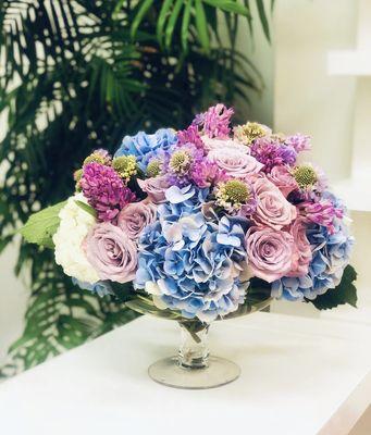 An arrangement of fresh hyacinth, Moody Blues roses, hydrangea and scabiosa designed inside of a footed vase.