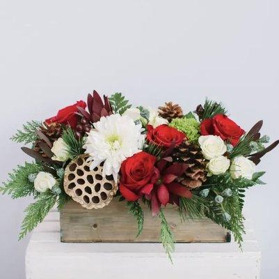 A classic holiday mix of red, green, white, and gold. This lush arrangement features red roses, winter greens, pine cones, and more.