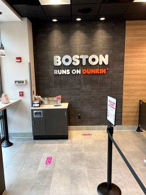 Dunkin' lobby, picturing social distancing stickers on the ground and a grey-stone wall with the lettering "BOSTON RUNS ON DUNKIN'"