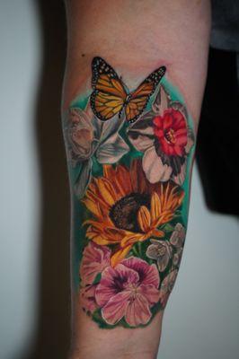 This vibrant tattoo features a monarch butterfly fluttering over colorful blooms, symbolizing beauty, transformation, and nature's grace.