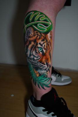 This vibrant tiger tattoo, framed by lush green leaves, captures the wild beauty and intense gaze of the jungle's most majestic predator.