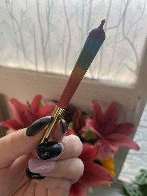 A rainbow joint held by a hand with long nails with flowers in the background. Taken at Little Green Pharmacy in Denver, CO.