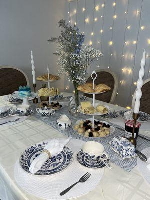 Blue and white tea party decorations. White 3 tiered holder with scones, sandwiches, and pastries from top to bottom. Floral centerpiece.