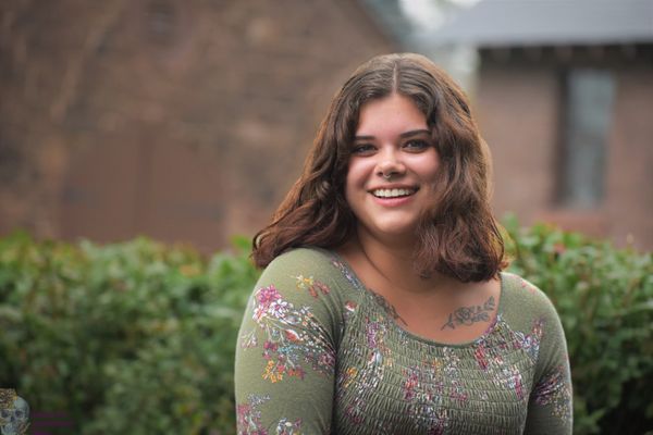 A girl smiling outside, in front of a building and bushes.